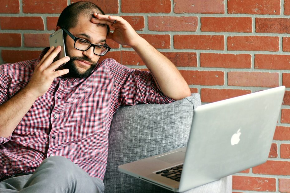 mężczyzna na tle ściany z cegieł. siedzi na kanapie rozmawiając przez telefon. obok leży otwarty laptop. mężczyzna spogląda na niego. drugą ręką podpiera głowę